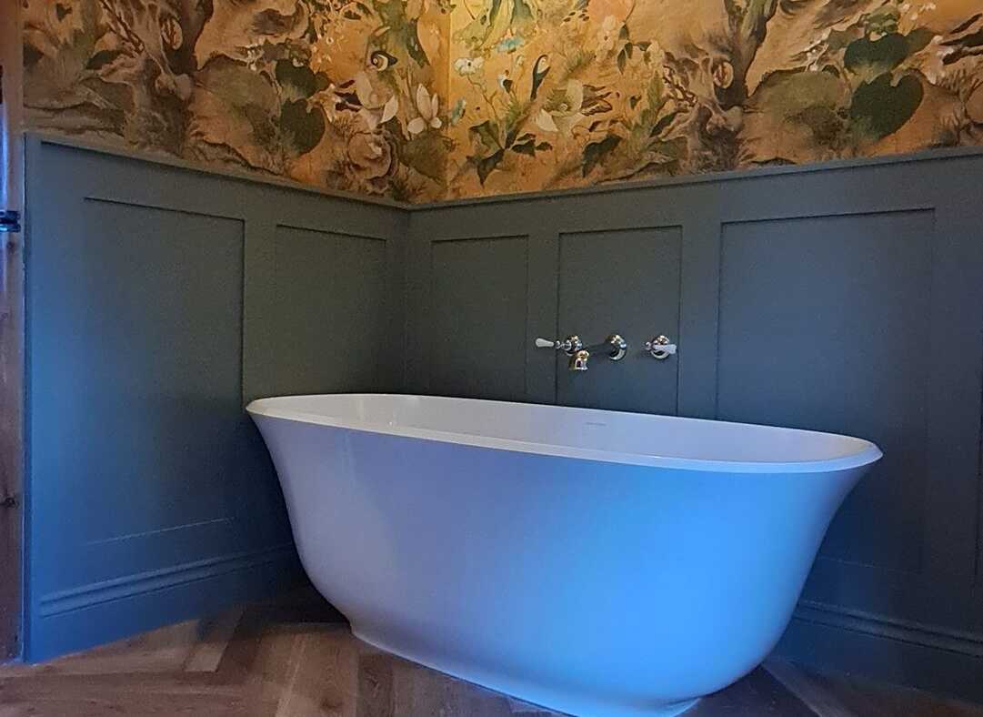 A freestanding bath against green panelling with floral wallpaper above