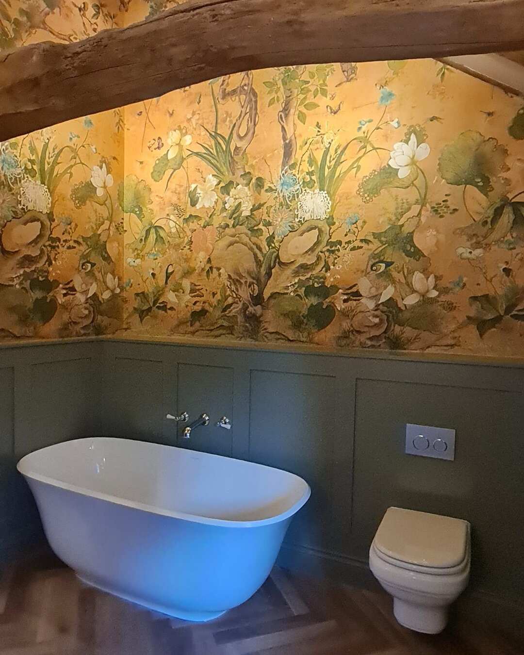 A freestanding bath against green panelling, with floral wallpaper and an old beam above