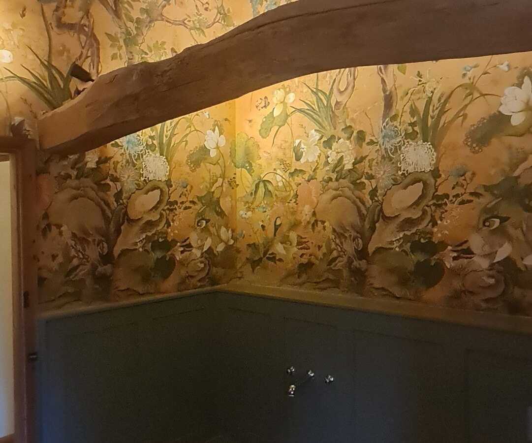 Floral wallpaper above painted panelling, under an old beam
